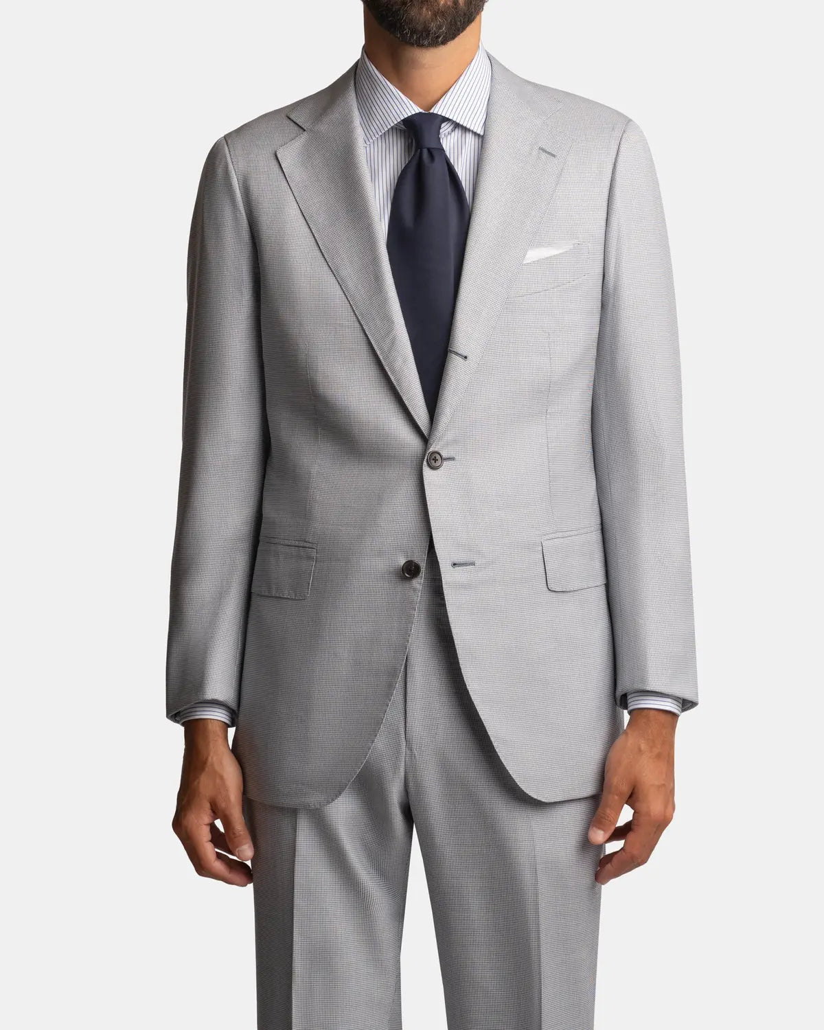 A man wears the Cavour Pale Blue Wool/Silk Puppytooth Suit with notch lapels, matching trousers, a white pocket square, a white striped shirt, and a dark navy tie. The image is cropped above his knees and his face is not visible. , available at Richard Gelding Mayfair