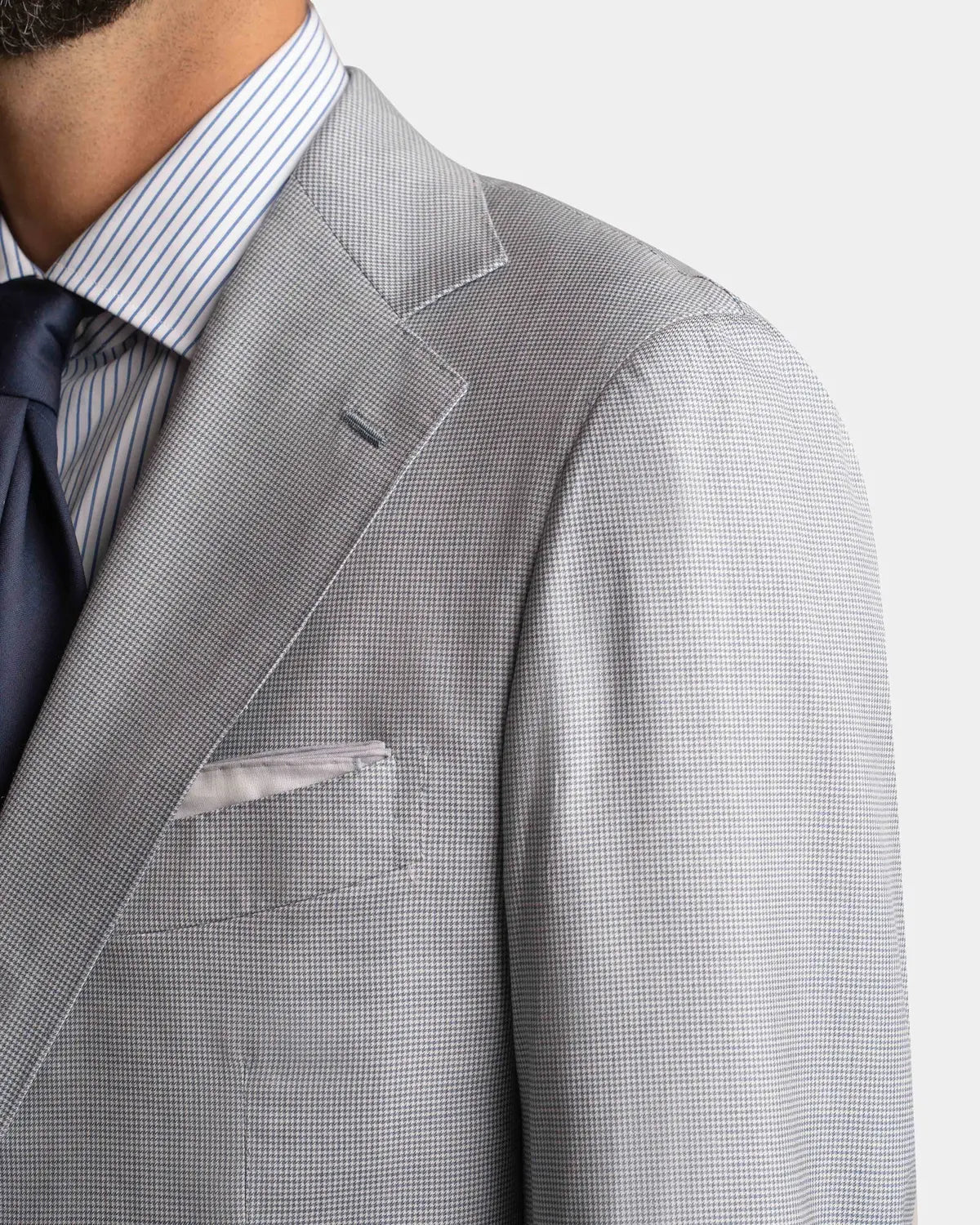 Close-up of a man in a Cavour Pale Blue Wool/Silk Puppytooth Suit jacket, paired with a white and blue striped shirt, dark navy tie, and light pocket square. Only his lower face and upper torso are visible. , available at Richard Gelding Mayfair