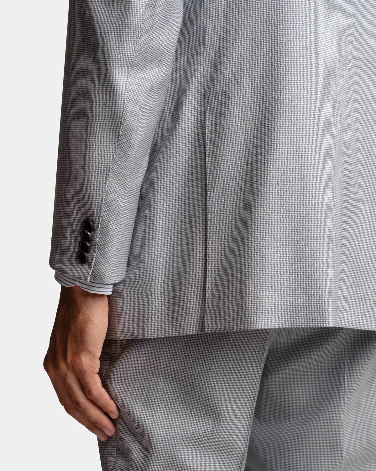 A close-up of a person wearing the Cavour Pale Blue Wool/Silk Puppytooth Suit from behind shows the left arm and side, with three dark buttons at the cuff. The suit's fine texture hints at its premium fabric, and their hand rests relaxed at their side. , available at Richard Gelding Mayfair