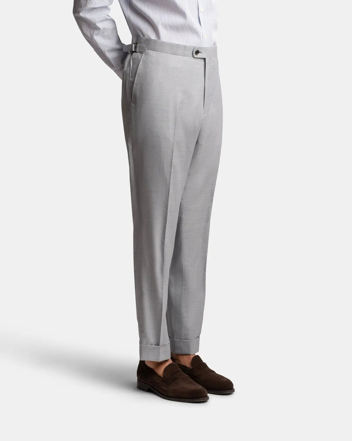 A cropped image shows only the lower torso and legs of a person in Cavour’s Pale Blue Wool/Silk Puppytooth Suit trousers, with side adjusters, brown loafers, and a tucked-in light blue and white striped shirt against a plain background. , available at Richard Gelding Mayfair