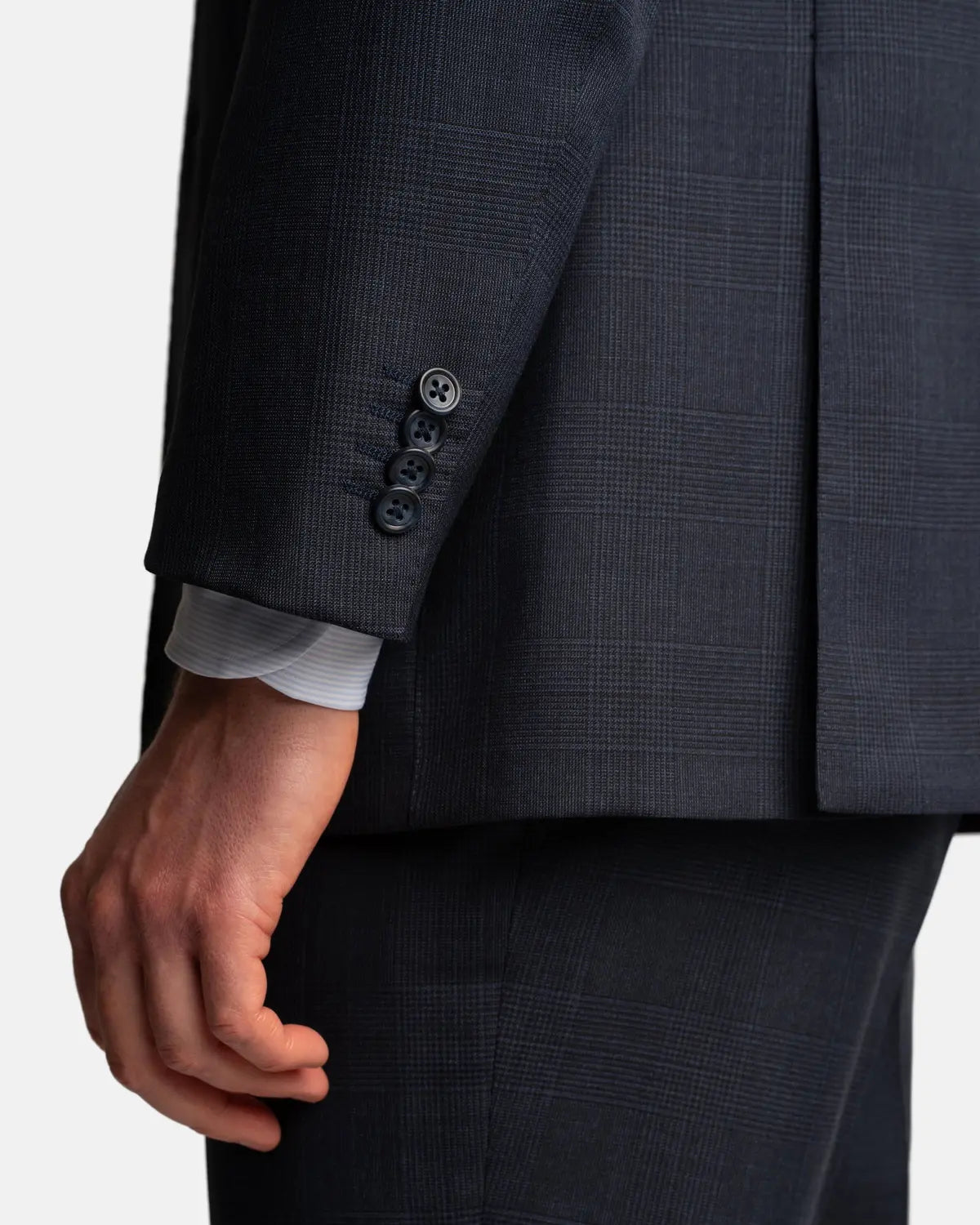 A close-up of a person’s left hand by their side, wearing the Cavour Navy Check Hightwist Wool Suit with four black sleeve buttons and a light gray striped shirt cuff peeking out. The background is plain white. , available at Richard Gelding Mayfair