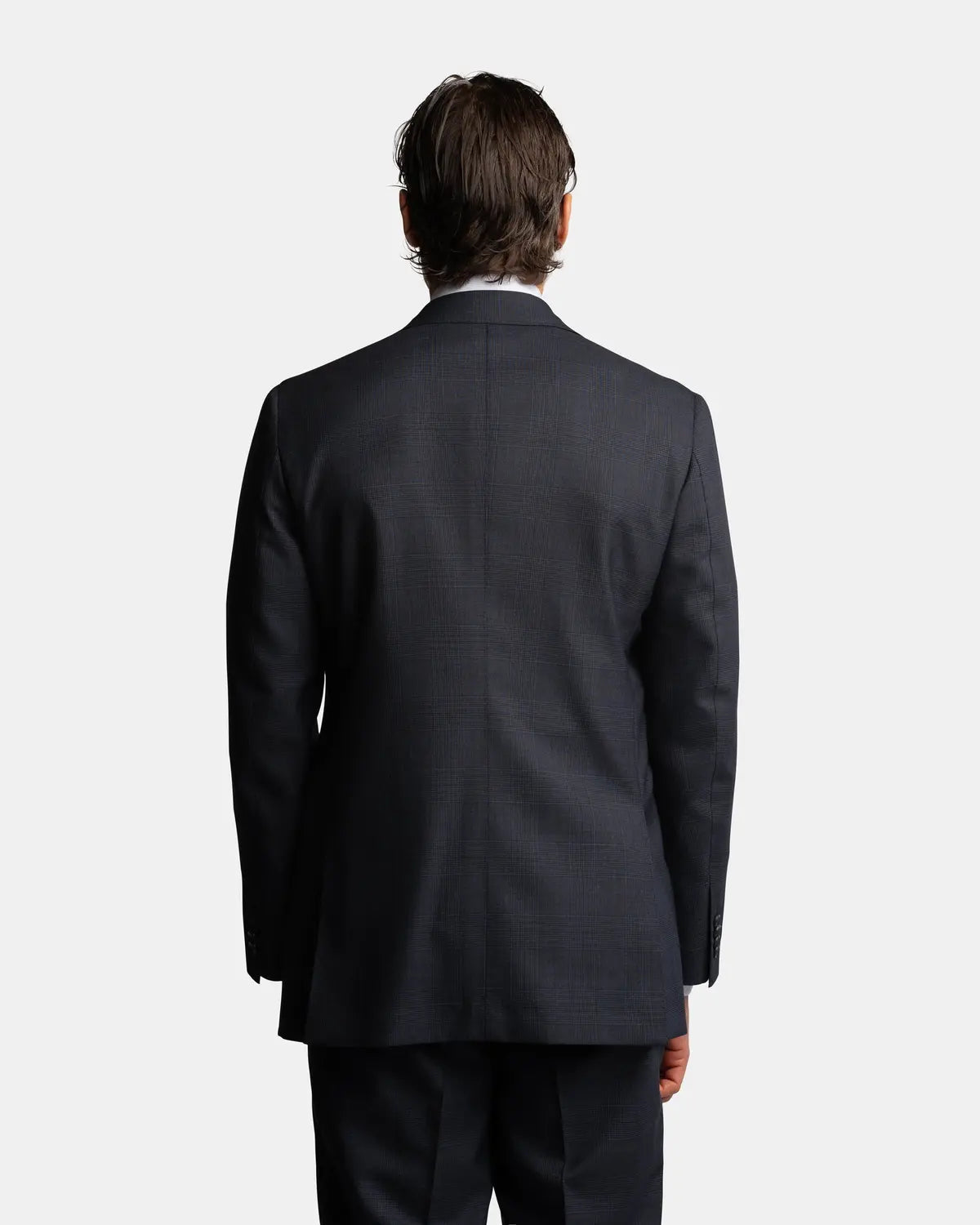 A man with short brown hair stands with his back to the camera, wearing the Cavour Navy Check Hightwist Wool Suit, perfect for business travel or formal occasions. The 100% wool suit is shown against a plain white background. , available at Richard Gelding Mayfair