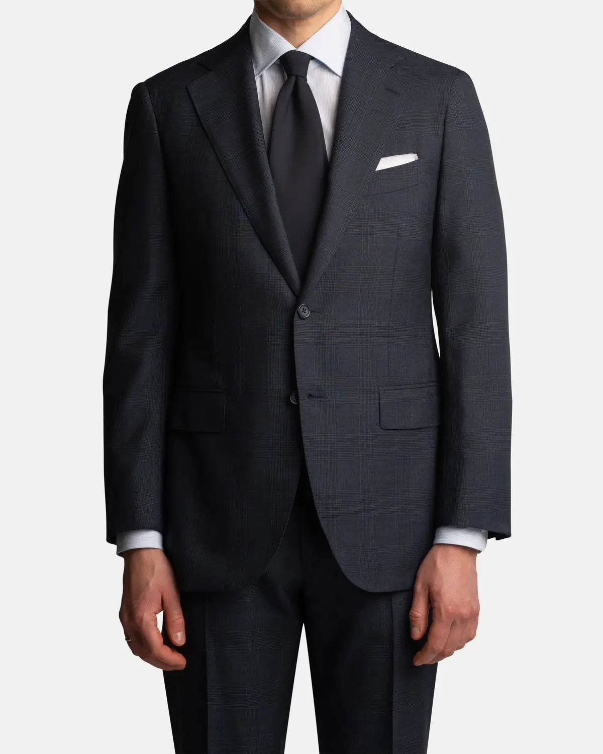 A man wears the Cavour Navy Check Hightwist Wool Suit—crafted from 100% wool with notch lapels—paired with a white dress shirt, black tie, and white pocket square. Hands at his sides; image cropped at neck and mid-thigh. , available at Richard Gelding Mayfair