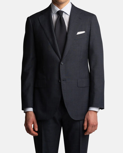 A man wears the Cavour Navy Check Hightwist Wool Suit—crafted from 100% wool with notch lapels—paired with a white dress shirt, black tie, and white pocket square. Hands at his sides; image cropped at neck and mid-thigh. , available at Richard Gelding Mayfair