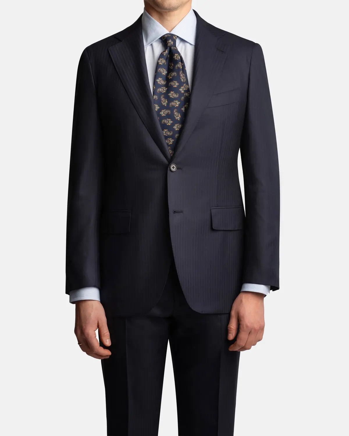 A man faces forward wearing the Cavour Navy Herringbone Super 150's Wool Suit, paired with a light blue shirt and navy tie. His head is slightly cropped out, hands at his sides, against a plain white background. , available at Richard Gelding Mayfair