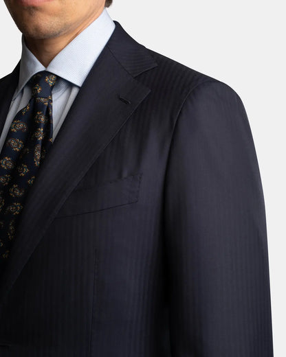 A man is shown from the chest up against a light background, wearing the Cavour Navy Herringbone Super 150's Wool Suit. Only the lower half of his face is visible. , available at Richard Gelding Mayfair