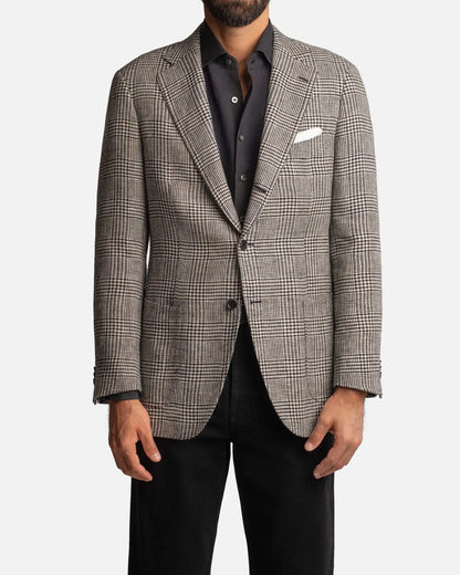 A man wears the Cavour Black/Grey Linen Glencheck Blazer, a single-breasted jacket with a classic notch lapel and two buttons, over a dark shirt with a white pocket square and black pants; only his torso and upper legs are visible. , available at Richard Gelding Mayfair