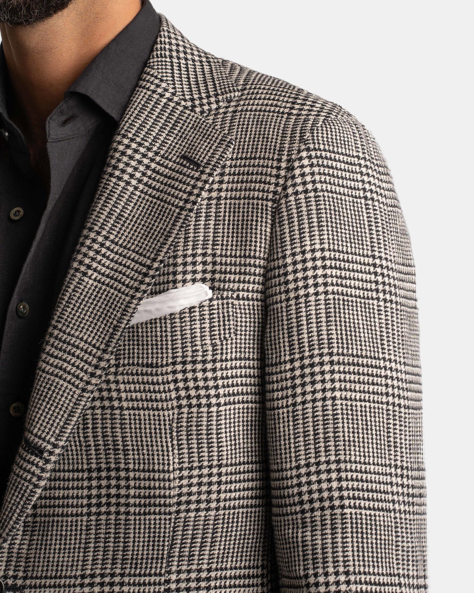 Close-up of a man in the Cavour Black/Grey Linen Glencheck Blazer, featuring a classic plaid in black, white, and grey. He wears a black shirt and displays a white pocket square; only the left side of his torso and face appear. , available at Richard Gelding Mayfair