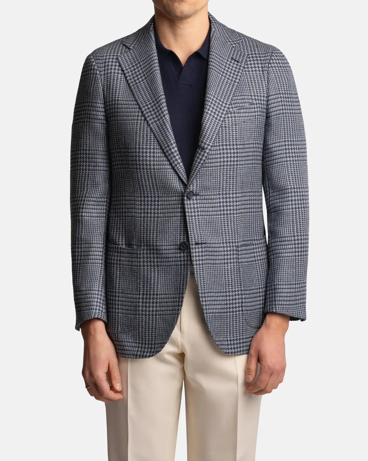 A man is shown from shoulders to mid-thigh, facing forward against a white background, wearing the Cavour Blue Glencheck Wool/Silk/Linen Blazer over a navy polo shirt and cream trousers, with his face partially cropped out. , available at Richard Gelding Mayfair