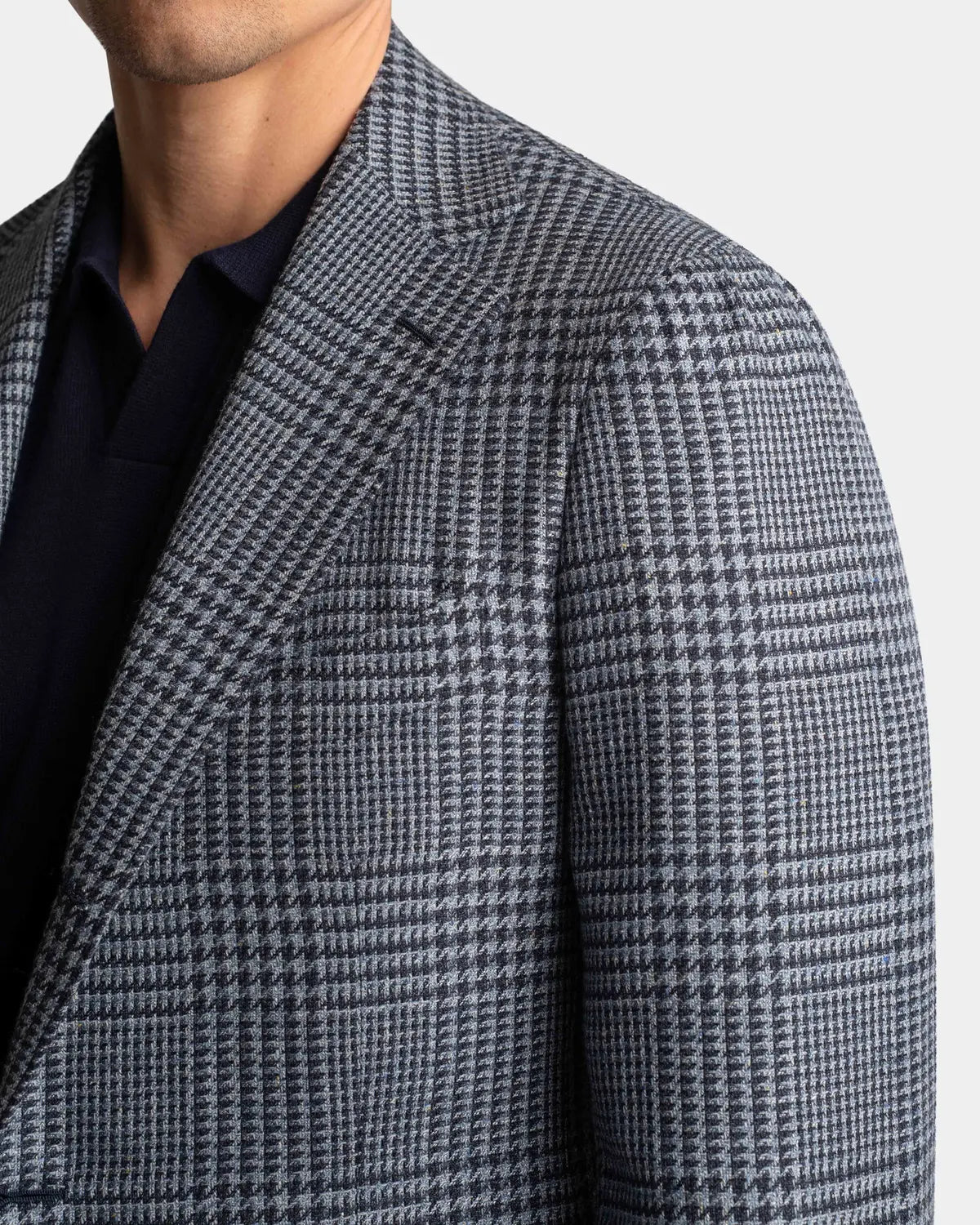 A close-up shows a man in a Cavour Blue Glencheck Wool/Silk/Linen Blazer over a dark navy collared shirt, with only his lower face and upper torso visible against a plain light background. , available at Richard Gelding Mayfair