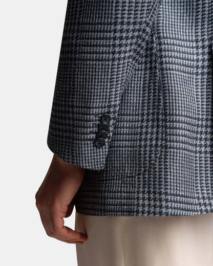 A close-up of a person’s arm in a Cavour Blue Glencheck Wool/Silk/Linen Blazer with two black cuff buttons, paired with light beige pants, against a plain white background. , available at Richard Gelding Mayfair
