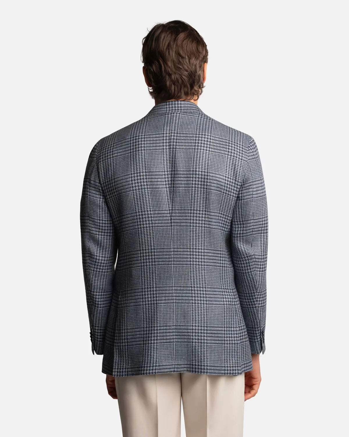 A man with medium-length brown hair is pictured from behind, wearing the Cavour Blue Glencheck Wool/Silk/Linen Blazer, paired with light trousers. The subtle check pattern stands out against the plain white background. , available at Richard Gelding Mayfair