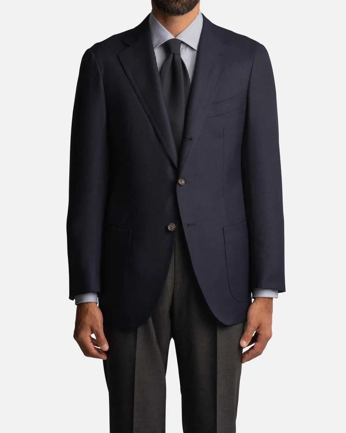 A man wears the Cavour Navy Hopsack Wool Blazer, featuring notch lapels, patch pockets, and two buttons, paired with a light blue shirt, dark tie, and dark gray trousers. He stands facing forward with hands at his sides; head partially out of frame. , available at Richard Gelding Mayfair