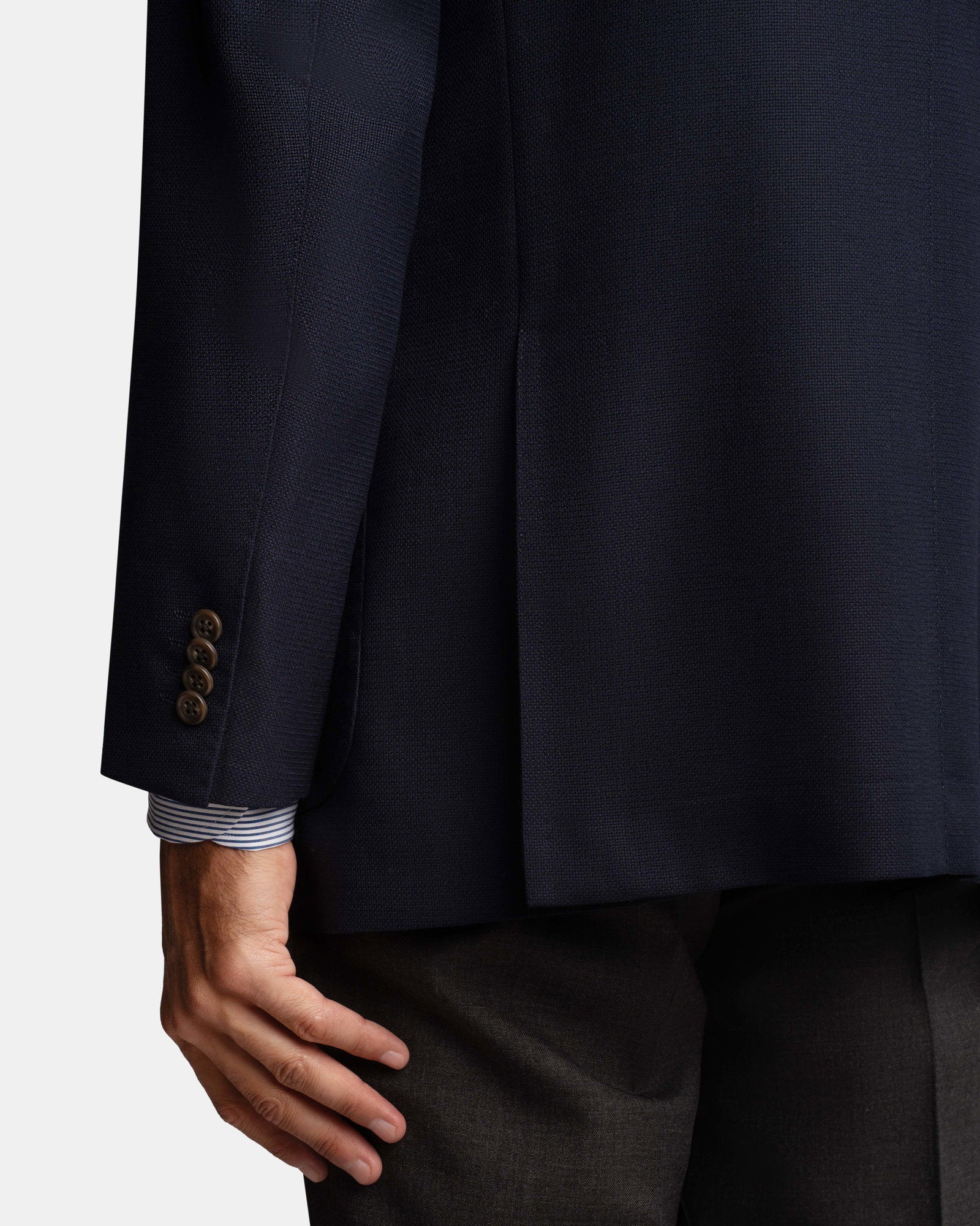 A close-up of a person's left hand by their side, wearing the Cavour Navy Hopsack Wool Blazer with four brown cuff buttons, a blue and white striped shirt cuff showing, and dark trousers against a plain light background. , available at Richard Gelding Mayfair