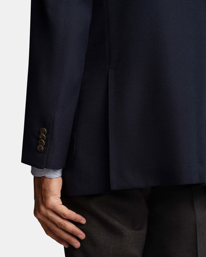 A close-up of a person's left hand by their side, wearing the Cavour Navy Hopsack Wool Blazer with four brown cuff buttons, a blue and white striped shirt cuff showing, and dark trousers against a plain light background. , available at Richard Gelding Mayfair