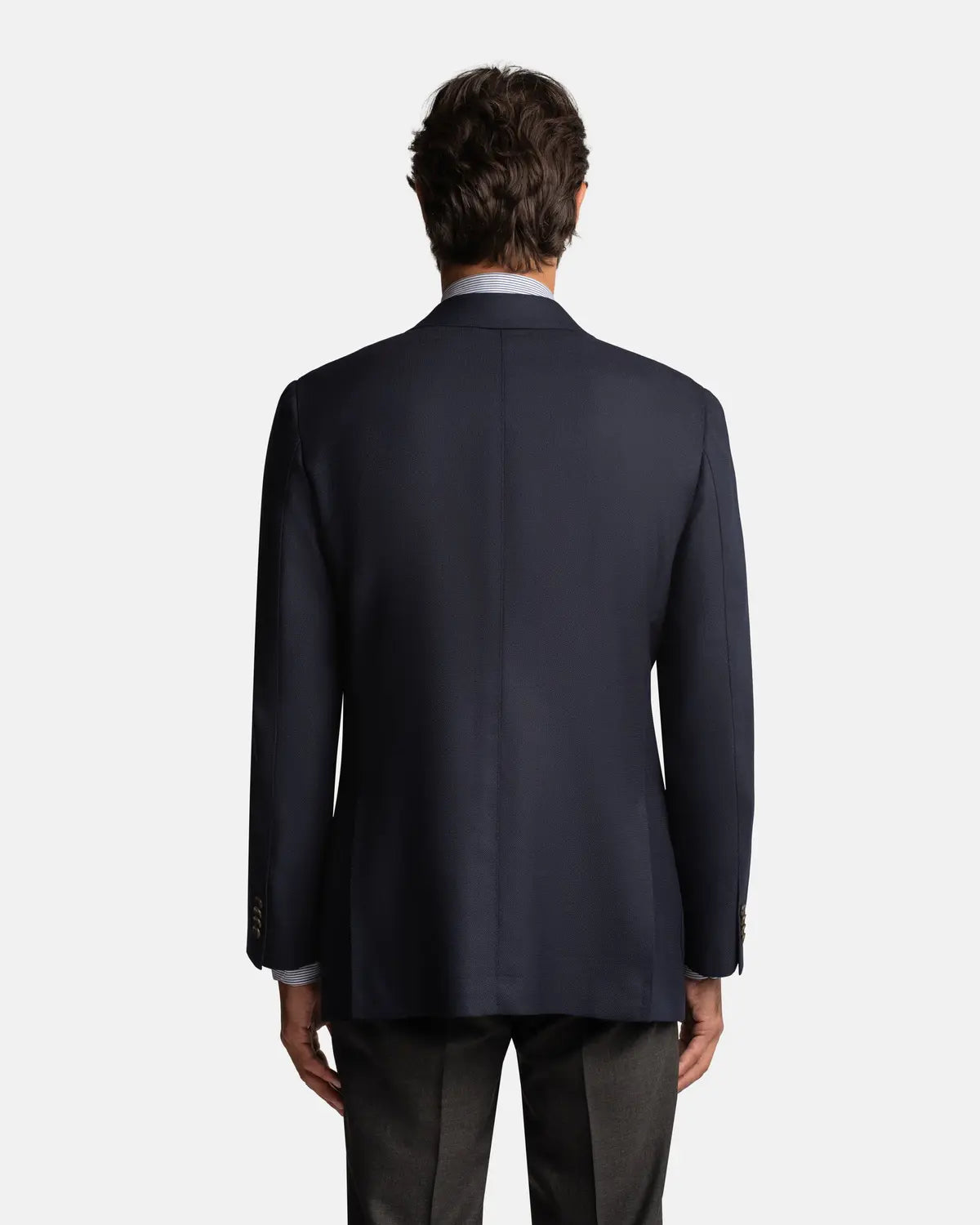 A man faces away from the camera wearing the Cavour Navy Hopsack Wool Blazer over a white shirt and dark trousers. His arms are relaxed by his sides, with a plain light gray background highlighting this versatile blazer. , available at Richard Gelding Mayfair