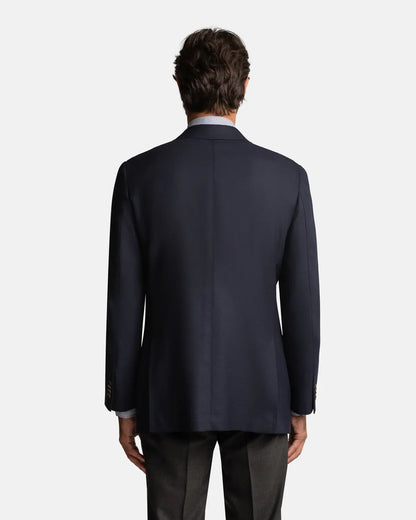 A man faces away from the camera wearing the Cavour Navy Hopsack Wool Blazer over a white shirt and dark trousers. His arms are relaxed by his sides, with a plain light gray background highlighting this versatile blazer. , available at Richard Gelding Mayfair