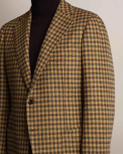 A Brown Shepherds Check Tweed Blazer by Cavour in a brown, green, and tan checkered pattern with notched lapels and two brown buttons, worn over a dark brown turtleneck sweater against a plain, light-colored background. , available at Richard Gelding Mayfair