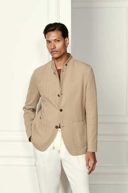 A person with short dark hair and a neutral expression stands indoors, wearing the Gran Sasso Beige Herringbone Cotton Blend Jacket over a brown shirt and white pants. The modern, minimalist style is highlighted by one hand casually in their pocket. , available at Richard Gelding Mayfair