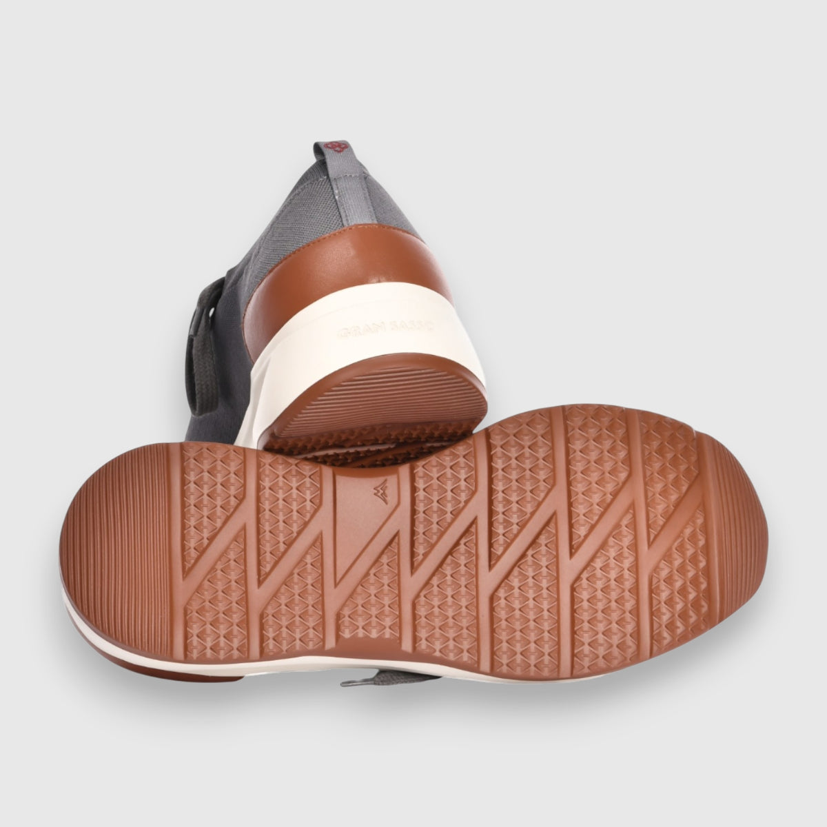 The Gran Sasso Grey Knitted Sneaker features a breathable knit upper in light gray with a black pull tab, gum-colored soles with geometric tread, and brown and white heels, shown from the back and bottom. , available at Richard Gelding Mayfair
