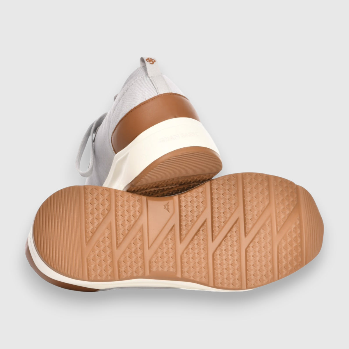 Gran Sasso’s Light Grey Knitted Sneakers feature white fabric uppers, tan brown heel accents, and light brown rubber soles. One slip-on sneaker stands upright to show the heel; the other displays its textured sole on a gray background. , available at Richard Gelding Mayfair