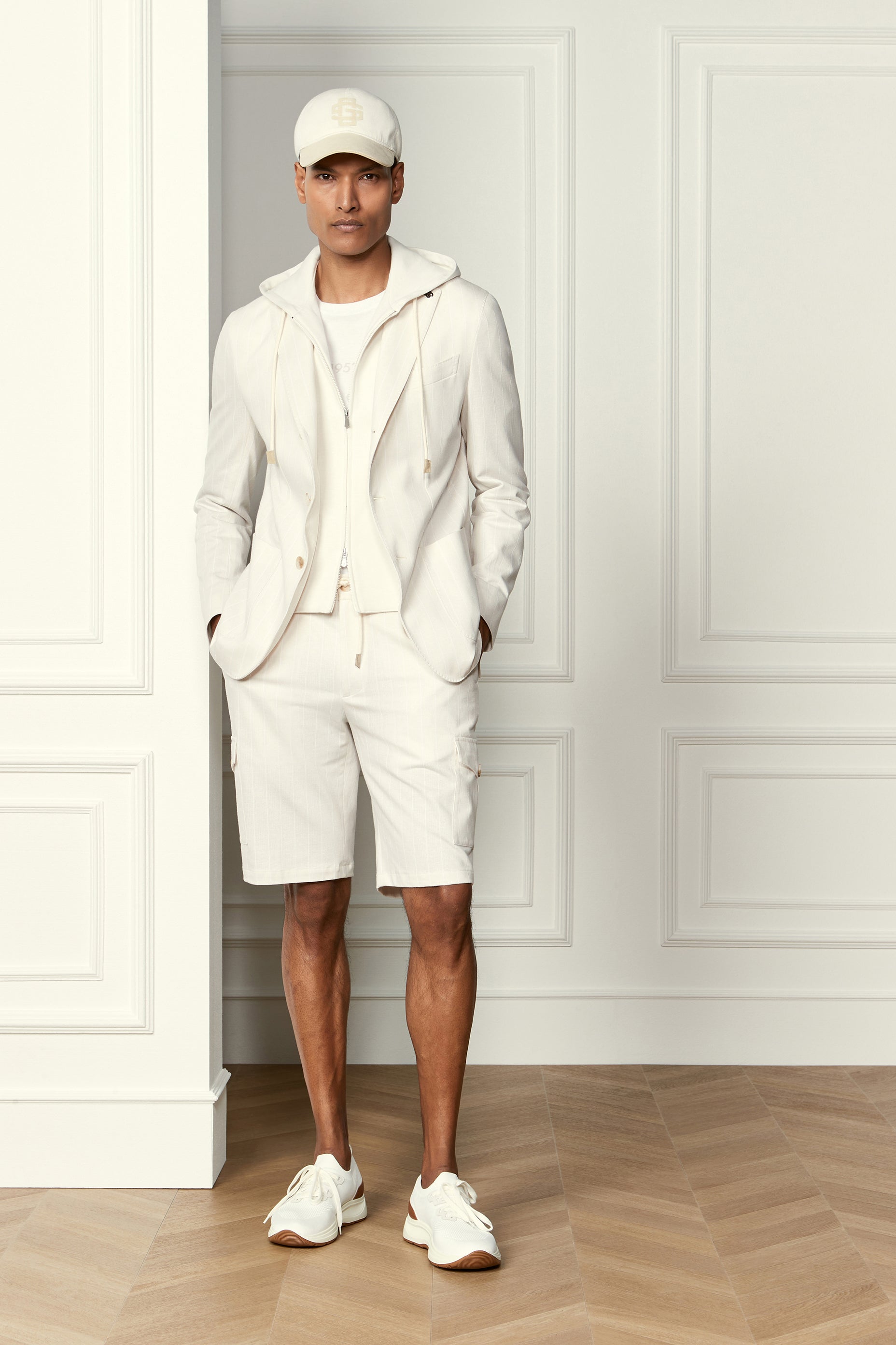 A man stands indoors against white paneled walls and a light wood floor, wearing Gran Sasso's White Knitted Sneakers with tan soles. He pairs them with a white cap, hoodie, blazer, and shorts, hands in his pockets as he faces the camera. , available at Richard Gelding Mayfair