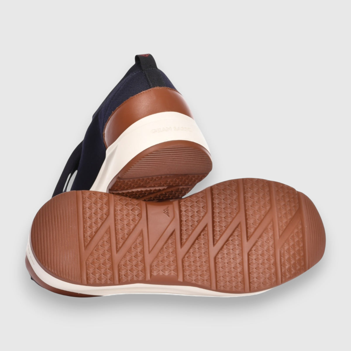 Close-up of Gran Sasso Navy Blue Knitted Sneakers: one upright, one on its side showing the rubber outsole with a zigzag and diamond pattern. Features navy uppers, brown heel accents, white midsoles. Light gray background. , available at Richard Gelding Mayfair