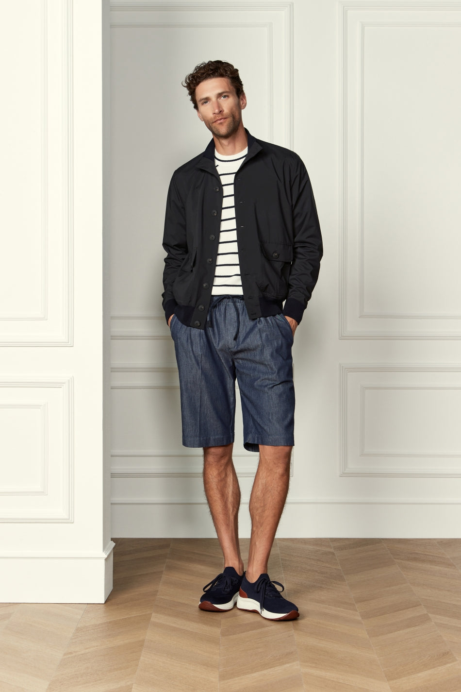 A man stands indoors against a white paneled wall, wearing navy blue knitted sneakers from Gran Sasso with white soles. He pairs them with a black jacket, a white and black striped shirt, blue drawstring shorts, and has short curly hair with a trimmed beard. , available at Richard Gelding Mayfair