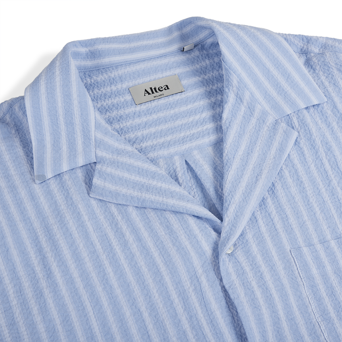 Close-up of the Altea Baby Blue Cotton Seersucker Cuban Shirt, featuring a textured vertical stripe pattern, notched collar, chest pocket, and visible “Altea Milano” label. Lightweight cotton makes it ideal for summer. , available at Richard Gelding Mayfair