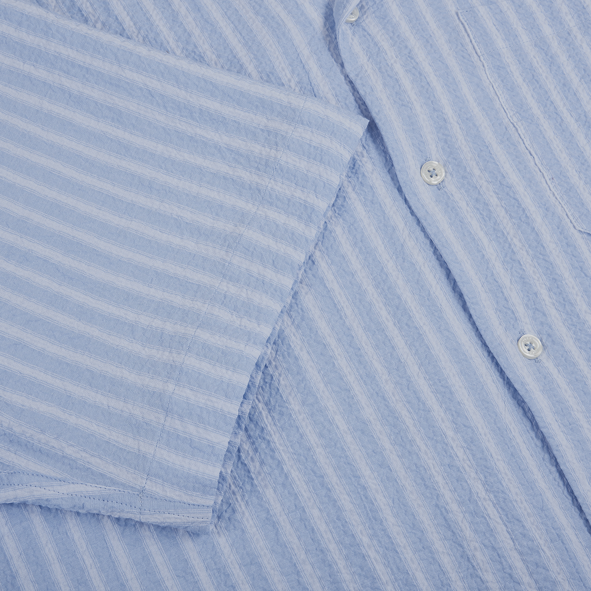 Close-up of the Altea Baby Blue Cotton Seersucker Cuban Shirt, featuring long sleeves and a vertical striped texture. The soft, slightly crinkled fabric makes it ideal for summer, with one sleeve partly covering the buttons and chest pocket. , available at Richard Gelding Mayfair