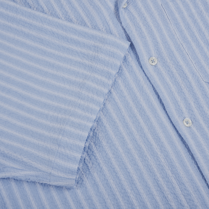Close-up of the Altea Baby Blue Cotton Seersucker Cuban Shirt, featuring long sleeves and a vertical striped texture. The soft, slightly crinkled fabric makes it ideal for summer, with one sleeve partly covering the buttons and chest pocket. , available at Richard Gelding Mayfair