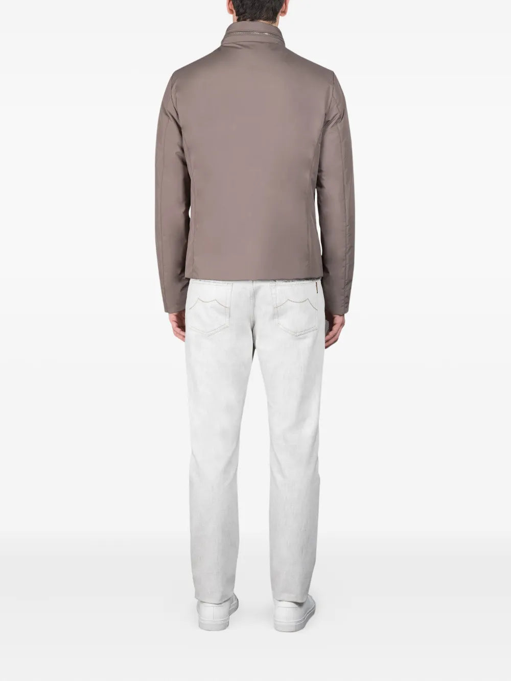 A man stands facing away from the camera against a plain white background, wearing the Moorer Ivory Avorio Nylon Alcide Blouson, light gray jeans, and white sneakers—a refined look exuding European sophistication. , available at Richard Gelding Mayfair