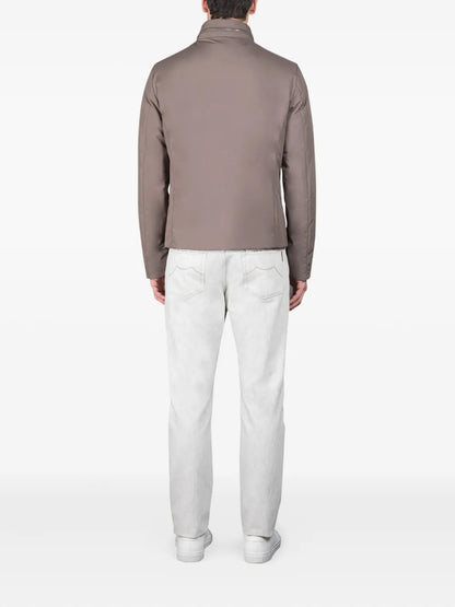 A man stands facing away from the camera against a plain white background, wearing the Moorer Ivory Avorio Nylon Alcide Blouson, light gray jeans, and white sneakers—a refined look exuding European sophistication. , available at Richard Gelding Mayfair