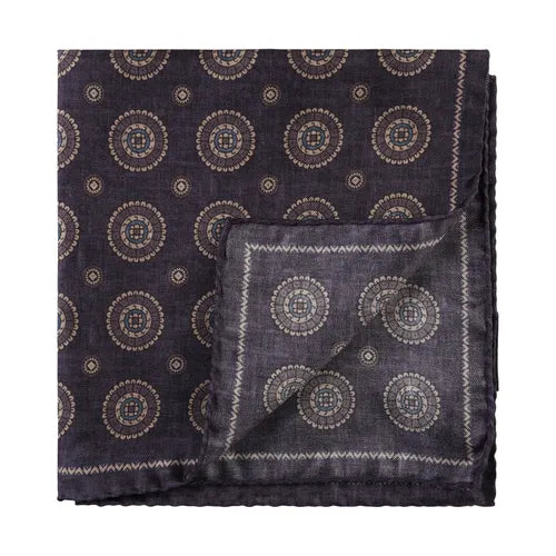The Amanda Christensen Medallion silk pocket square features a folded design in dark purple with a beige and light brown circular geometric pattern and a distinctive zigzag border. One corner is flipped to reveal the reverse side. , available at Richard Gelding Mayfair
