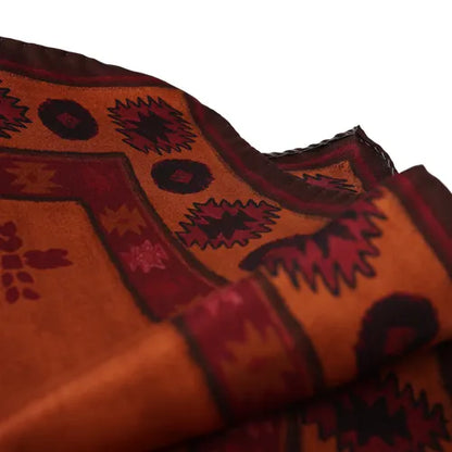 A close-up of the Amanda Christensen Silk pocket square with an Inca inspired pattern, showcasing geometric shapes in deep red, maroon, and black on a burnt orange background. The fabric appears soft with a visible, slightly wavy border and intricate, repeating designs. The background is plain white. , available at Richard Gelding Mayfair