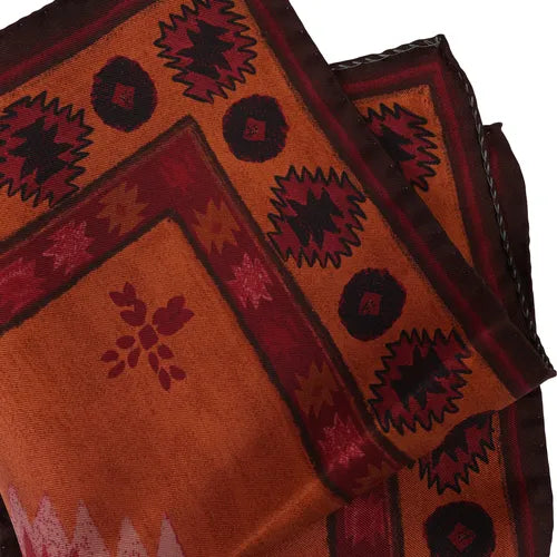 A silk pocket square by Amanda Christensen, featuring an Inca-inspired pattern in burnt orange and maroon with black and dark red geometric and floral motifs, including diamond and starburst designs along the borders. The printed fabric showcases a traditional, handcrafted appearance. , available at Richard Gelding Mayfair