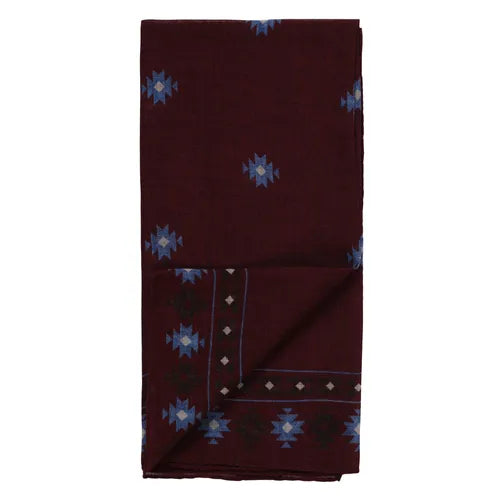 The Amanda Christensen Wool bandana features an Inca-inspired pattern in maroon, adorned with blue geometric designs and a border of small white diamond shapes and blue motifs, displayed against a plain background. , available at Richard Gelding Mayfair