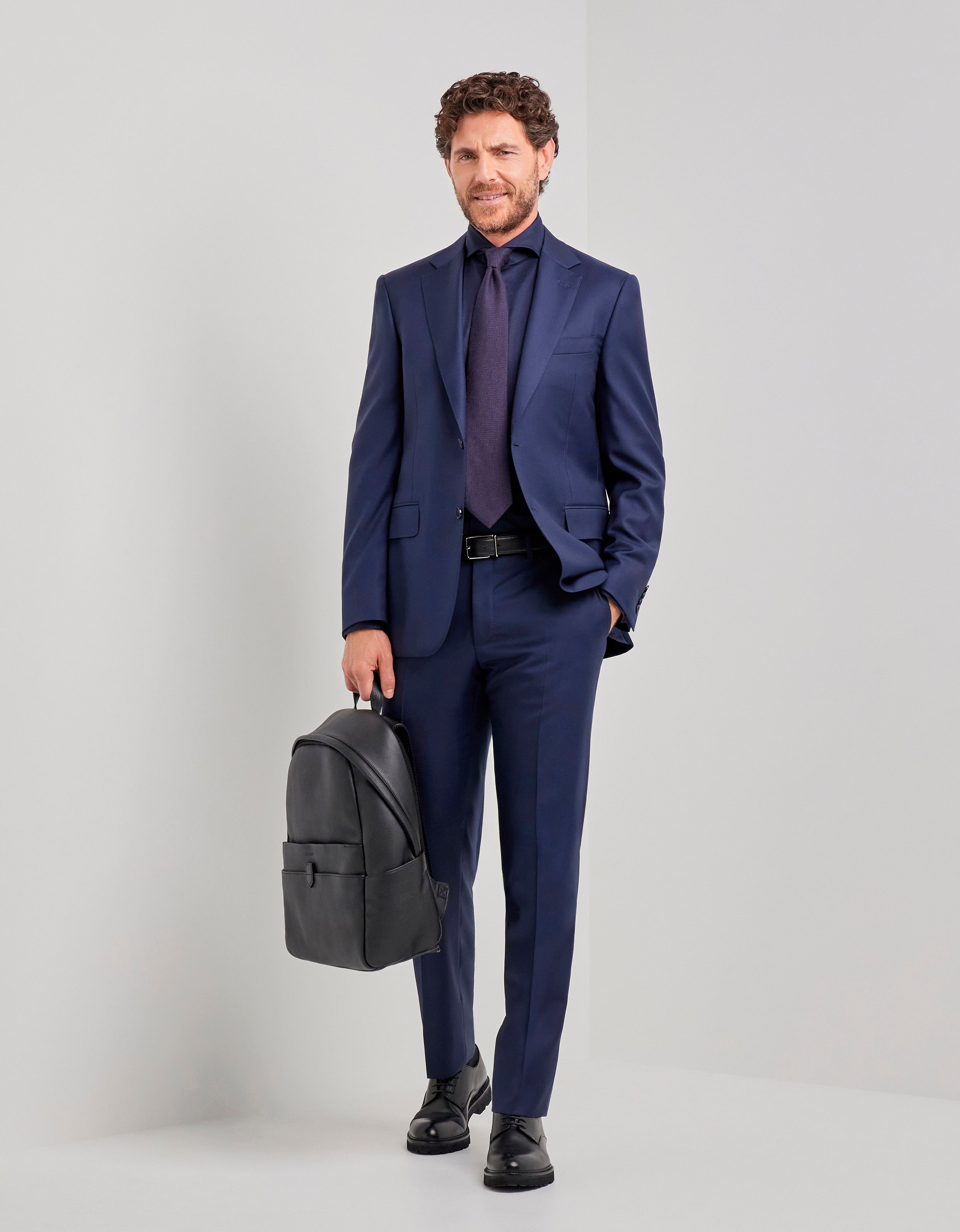 A man with curly hair wears the Canali Blue Wool Twill Single Breasted Suit with a dark shirt and matching tie. He stands against a light gray background, holding a black leather backpack in his left hand, looking relaxed and confident. , available at Richard Gelding Mayfair