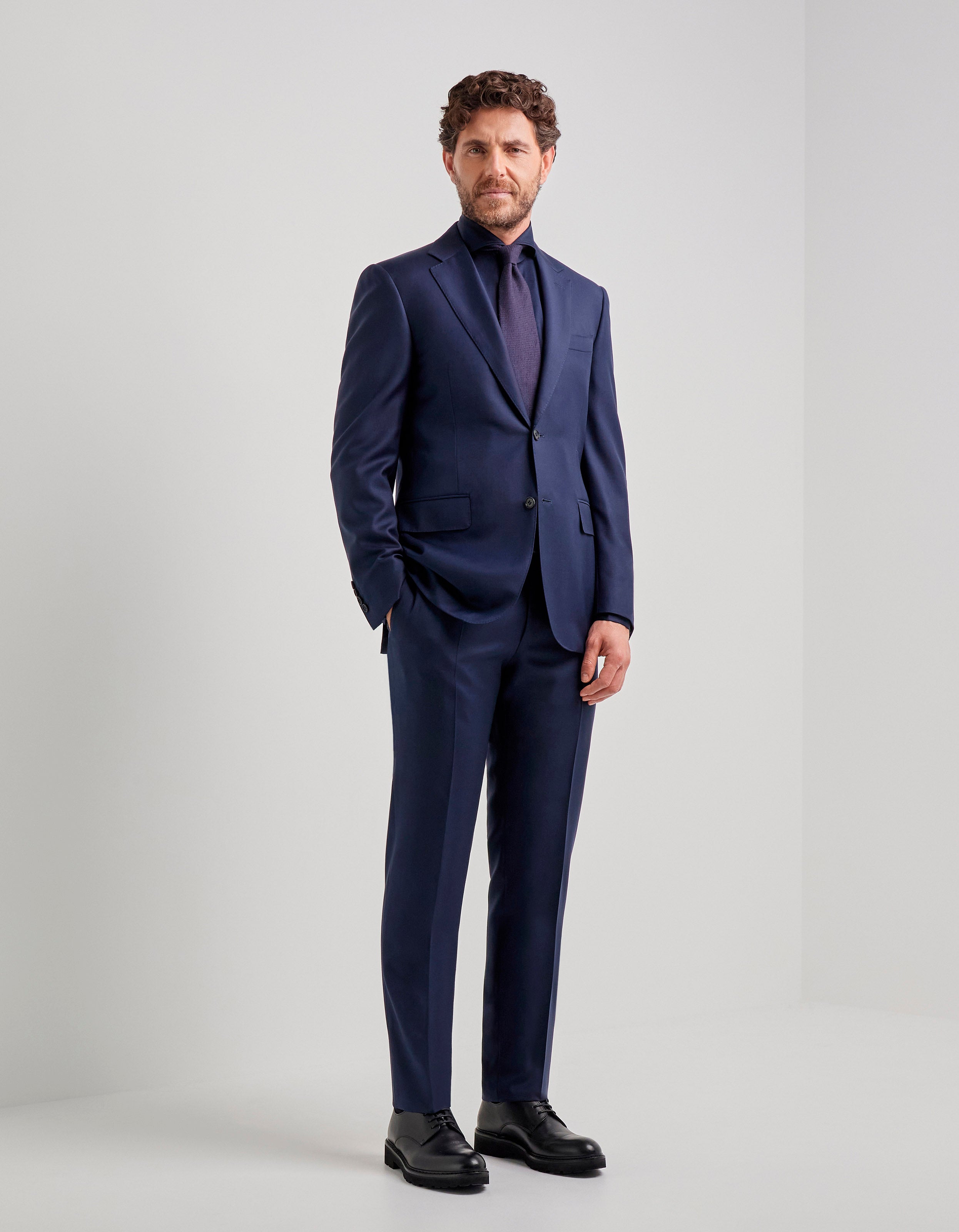 A man with short curly brown hair and a beard stands confidently in a plain white room, wearing the Canali Blue Wool Twill Single Breasted Suit with matching shirt and tie, black dress shoes, and one hand in his pocket. , available at Richard Gelding Mayfair