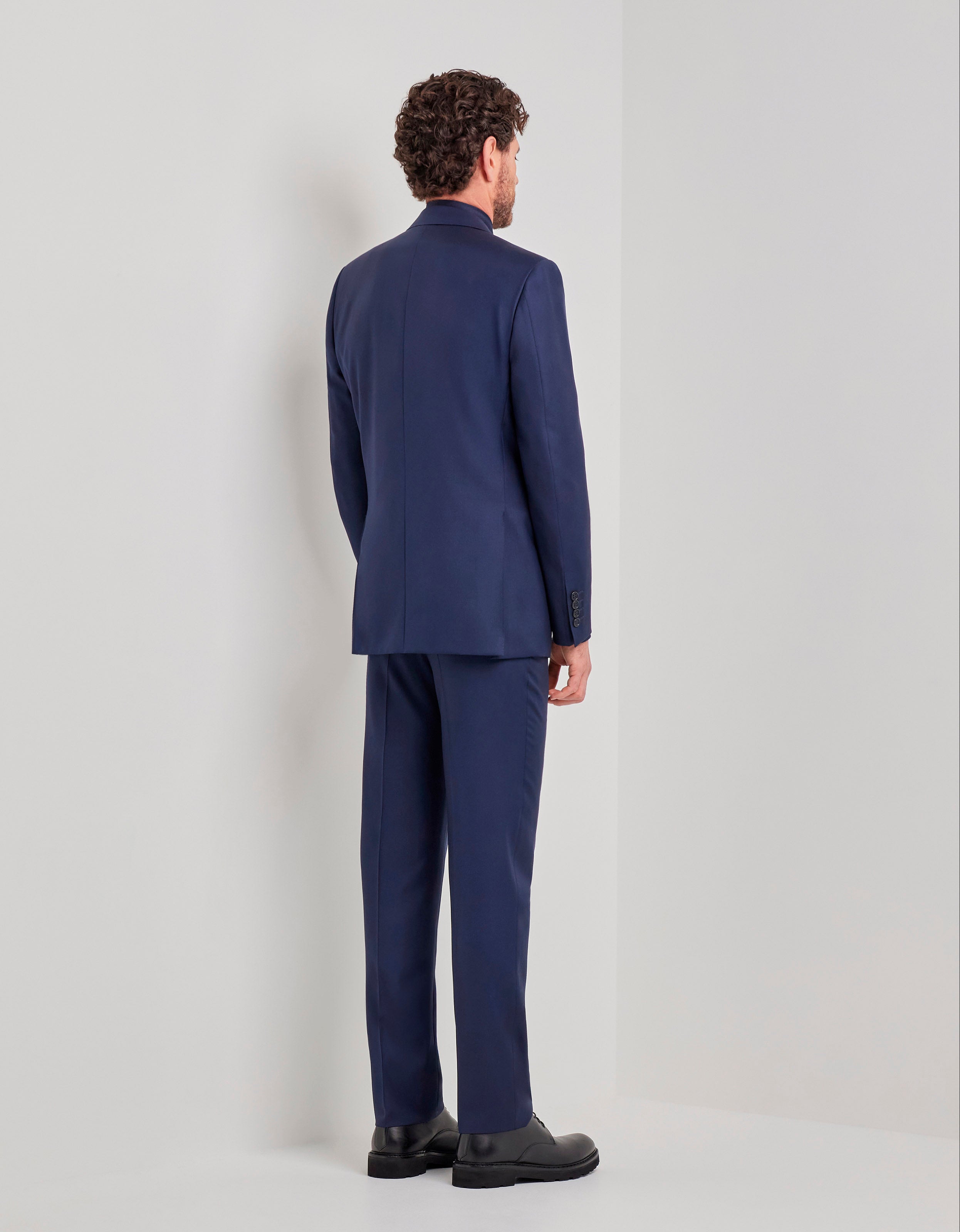 A man with curly brown hair, dressed in the Canali Blue Wool Twill Single Breasted Suit, stands with his back to the camera facing a white wall in a minimalist, light-colored room, his arms resting naturally by his sides. , available at Richard Gelding Mayfair