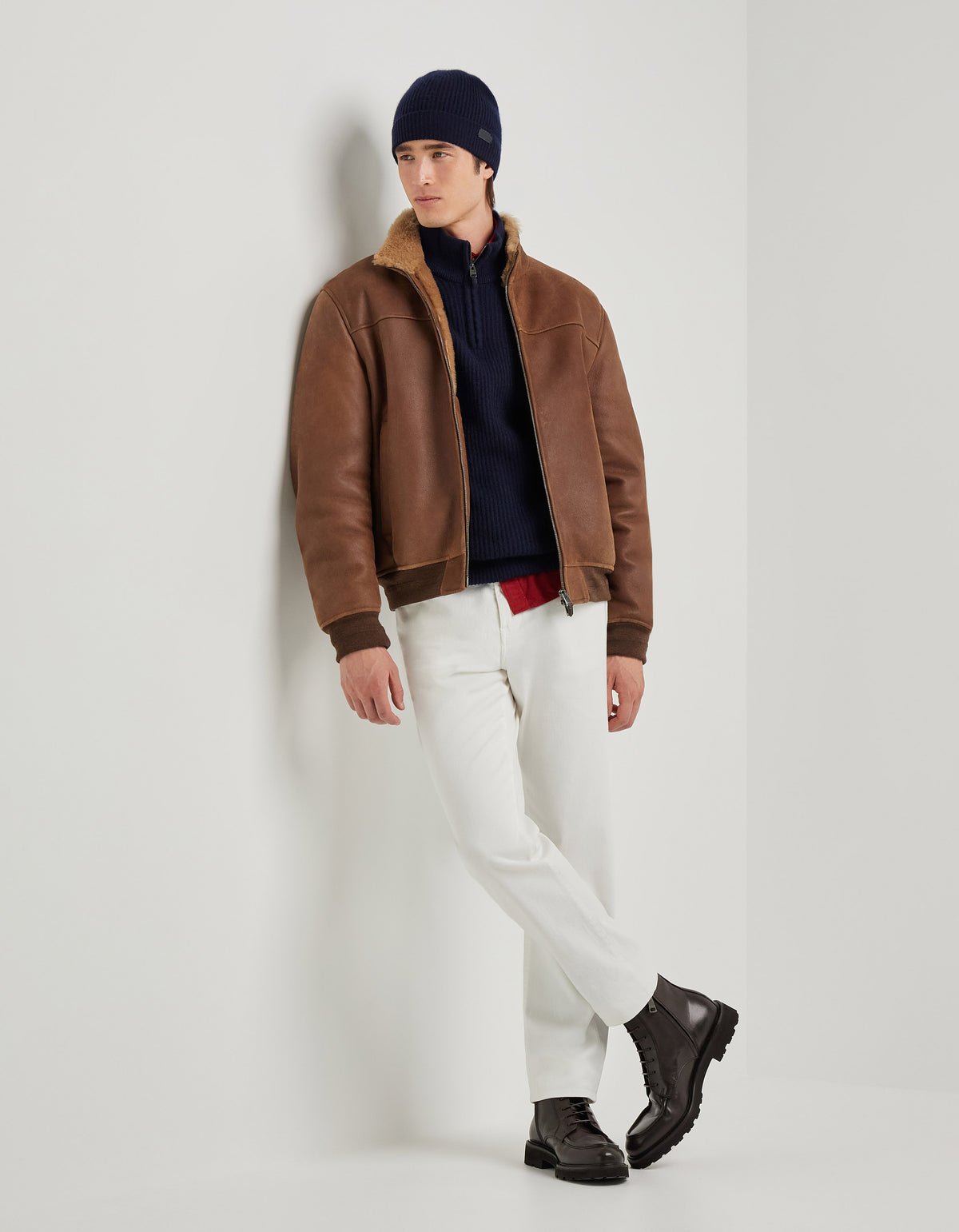 A man wearing the Canali Brown Merino Shearling Lambskin Suede Blouson, layered over a navy sweater and red shirt, pairs it with white pants, black boots, and a navy beanie as he leans against a white wall with arms relaxed and ankles crossed, gazing off to the side with a neutral expression in a minimal studio setting. , available at Richard Gelding Mayfair