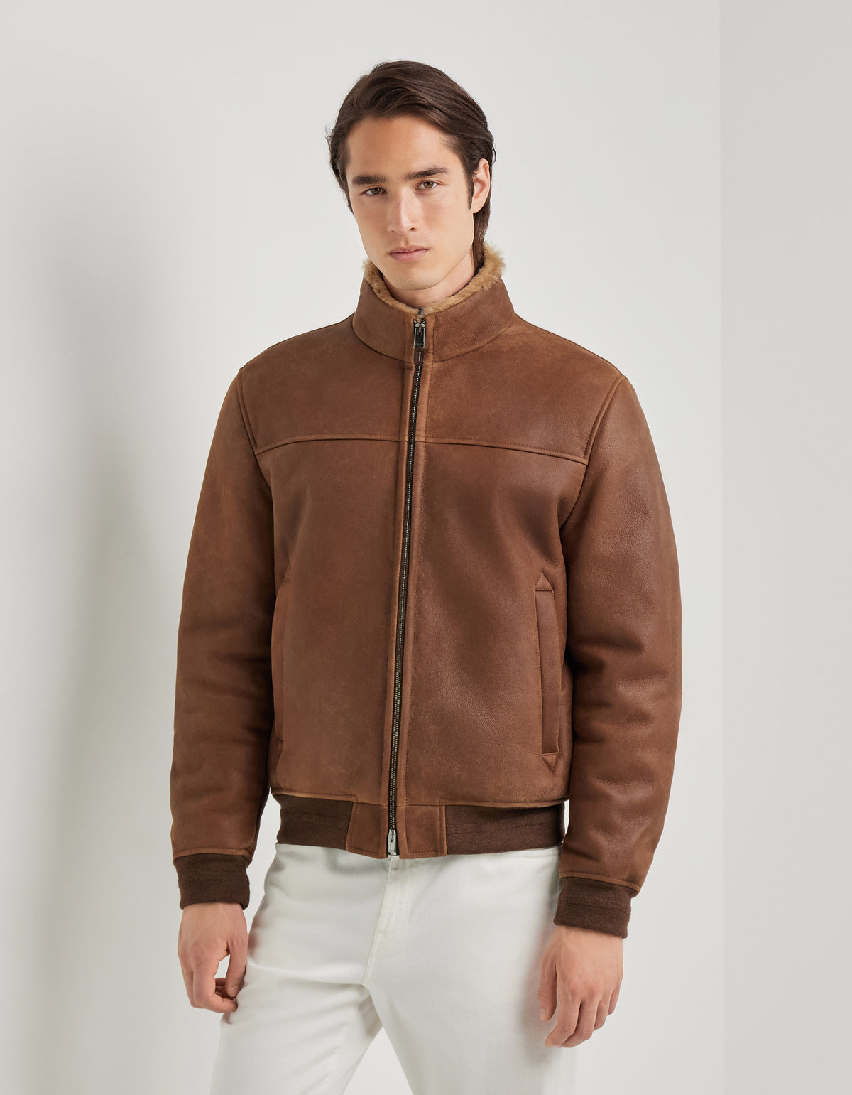 A person with long dark hair stands against a plain white wall, wearing a Brown Merino Shearling Lambskin Suede Blouson by Canali. The blouson features ribbed cuffs and hem with a zipper closure, paired with white pants, as the person poses with a neutral expression. , available at Richard Gelding Mayfair