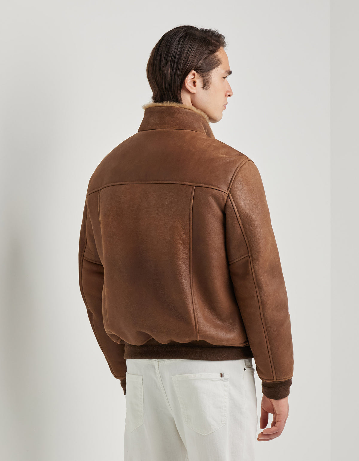 A person stands facing away, wearing the Canali Brown Merino Shearling Lambskin Suede Blouson with a raised collar and white pants. The blouson showcases visible stitching and a slightly relaxed fit. The background is plain white, and the person’s dark hair is neatly combed. , available at Richard Gelding Mayfair