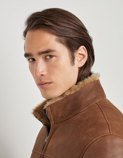 A young man with fair skin and straight brown hair looks at the camera with a neutral expression. He is wearing a Canali Brown Merino Shearling Lambskin Suede Blouson, crafted from luxurious lambskin suede and featuring a tan fur collar. The background is plain and light-colored. , available at Richard Gelding Mayfair