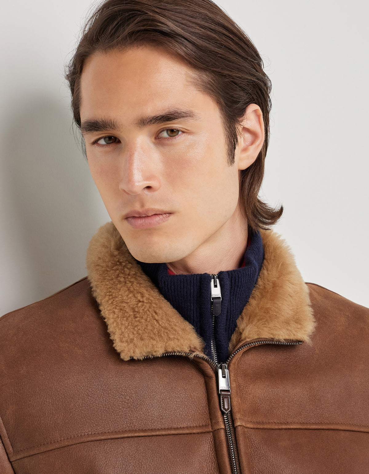 A young man with straight, brown hair wears Canali’s Brown Merino Shearling Lambskin Suede Blouson—a luxurious brown suede jacket featuring a tan fur collar—layered over a navy-blue zip-up sweater. He gazes seriously at the camera against a plain light background. , available at Richard Gelding Mayfair