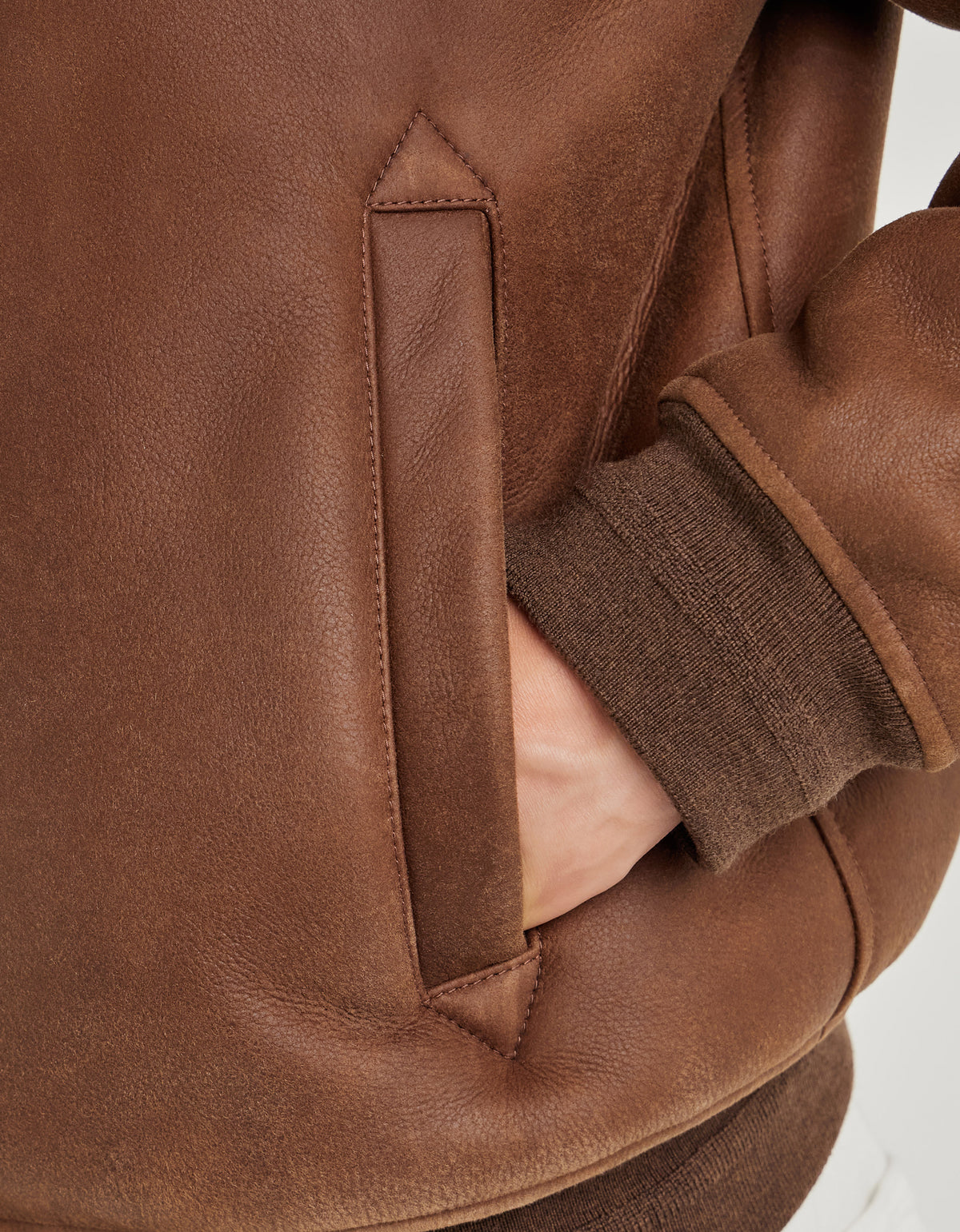A close-up of a person wearing the Canali Brown Merino Shearling Lambskin Suede Blouson with their hand in the jacket pocket. The sleeve is finished with ribbed brown knit fabric at the cuff, and the merino shearling suede has a soft, slightly textured appearance. The background is plain and neutral. , available at Richard Gelding Mayfair