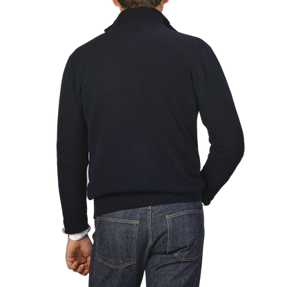 The image shows the back view of a person wearing a dark turtleneck sweater and blue jeans against a plain background, subtly hinting at the quality craftsmanship of an Altea Navy Blue Shetland Wool Button Cardigan.