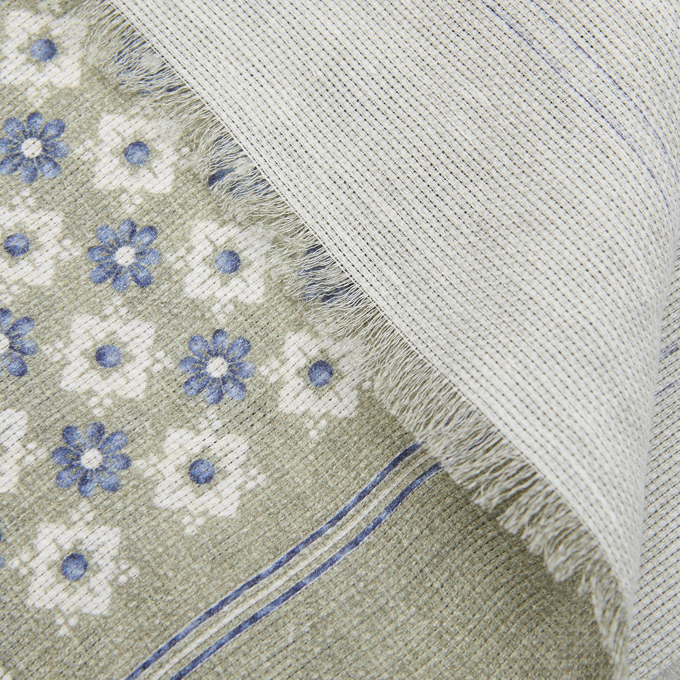 Close-up of an Amanda Christensen Olive Green Floral Printed Linen Cotton Scarf peeking under a solid gray fabric with frayed edges.