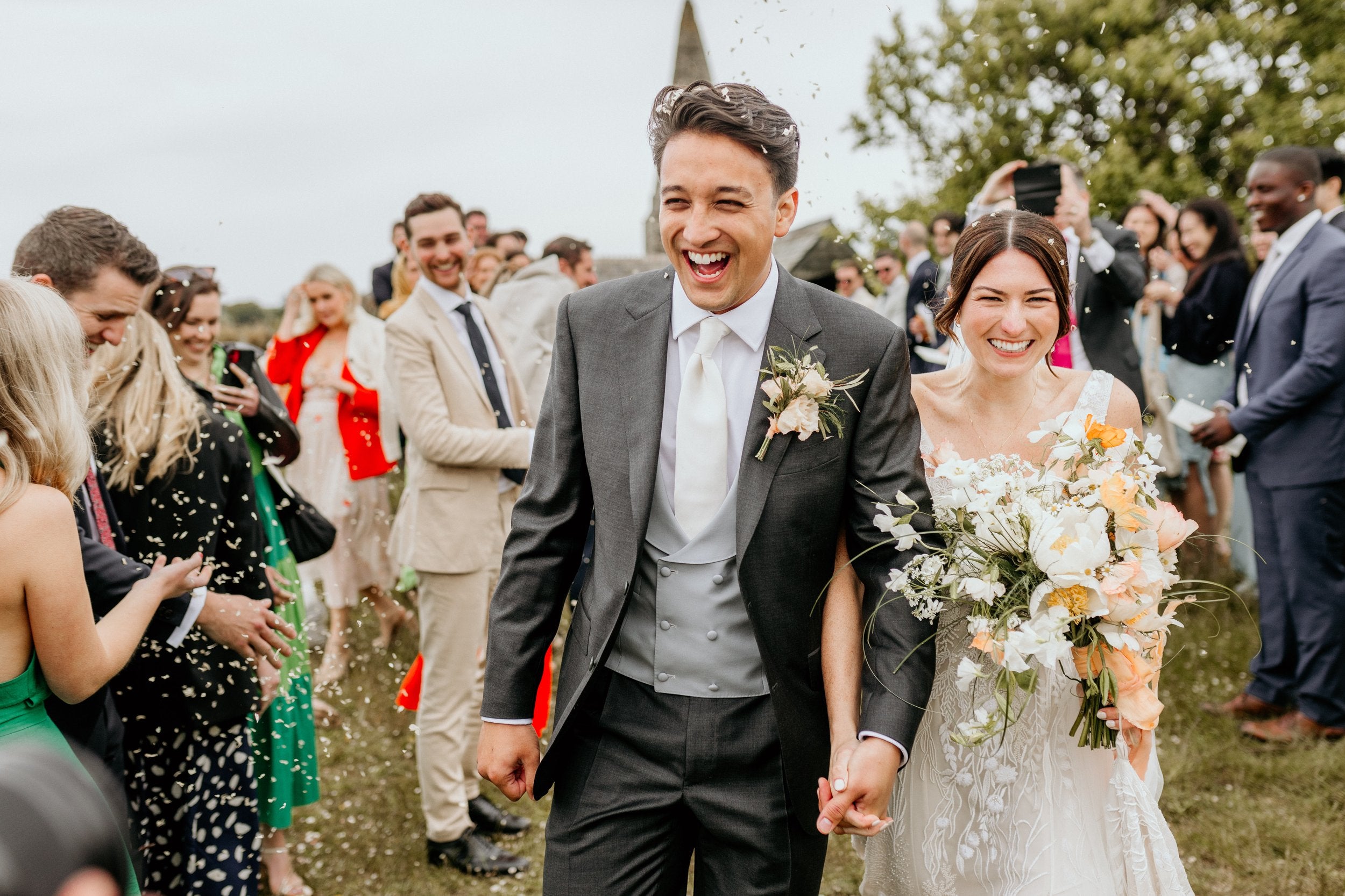 A joyful bride and groom walk hand in hand outdoors, smiling as guests throw confetti. The groom wears a gray suit and the bride holds a bouquet of white and peach flowers. People in festive attire celebrate around them on a grassy area. , available at Richard Gelding Mayfair
