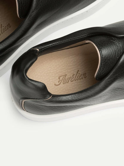 Close-up of two black grain leather slip-on sneakers by Aurélien, featuring white soles and tan inner lining. "Aurélien" is embossed in gold on the insole, showcasing these luxury shoes against a light neutral background. , available at Richard Gelding Mayfair